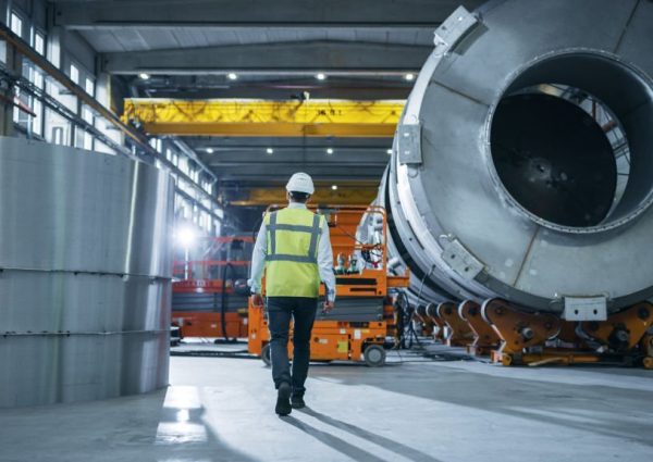 Following Shot of Heavy Industry Engineers Walking Through Manufacturing Factory. In the Background Professionals Working on Construction of Oil, Gas and Fuel Pipeline Transportation Products